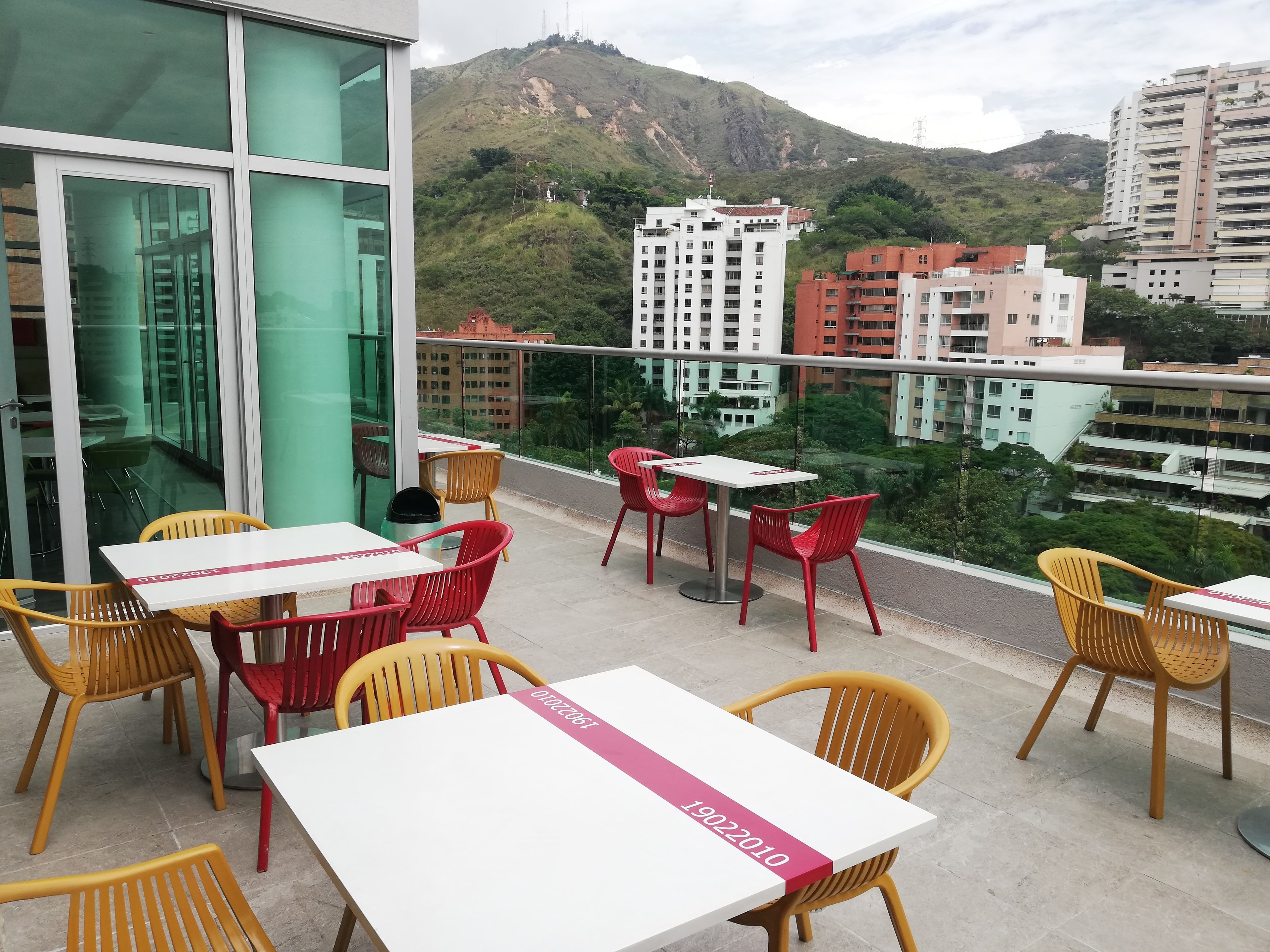 A balcony with tables and chairs, overlooking a cityscape with buildings and a hill.