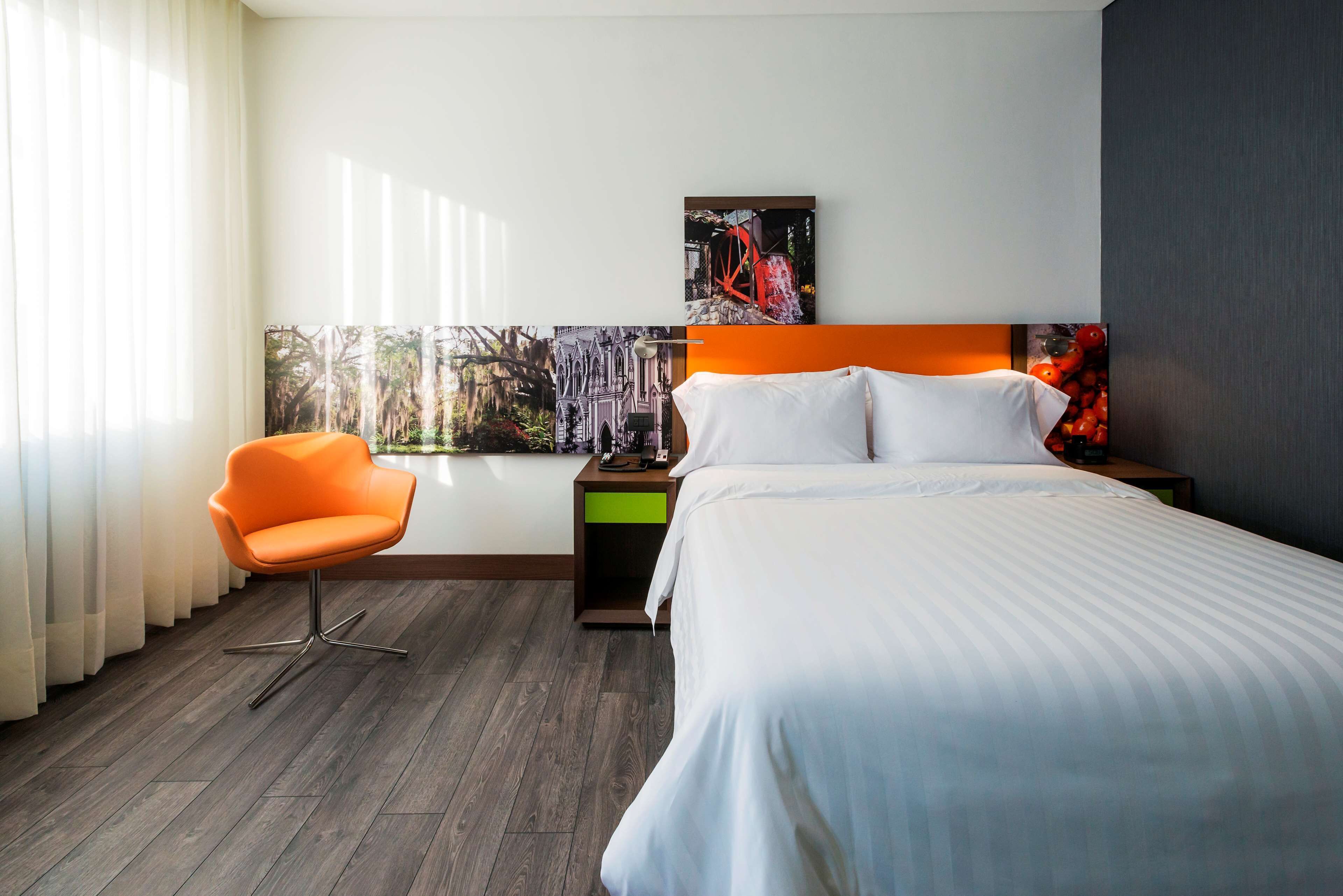 Una habitación de hotel con cama, una silla, una mesita de noche y un ventanal con cortinas.