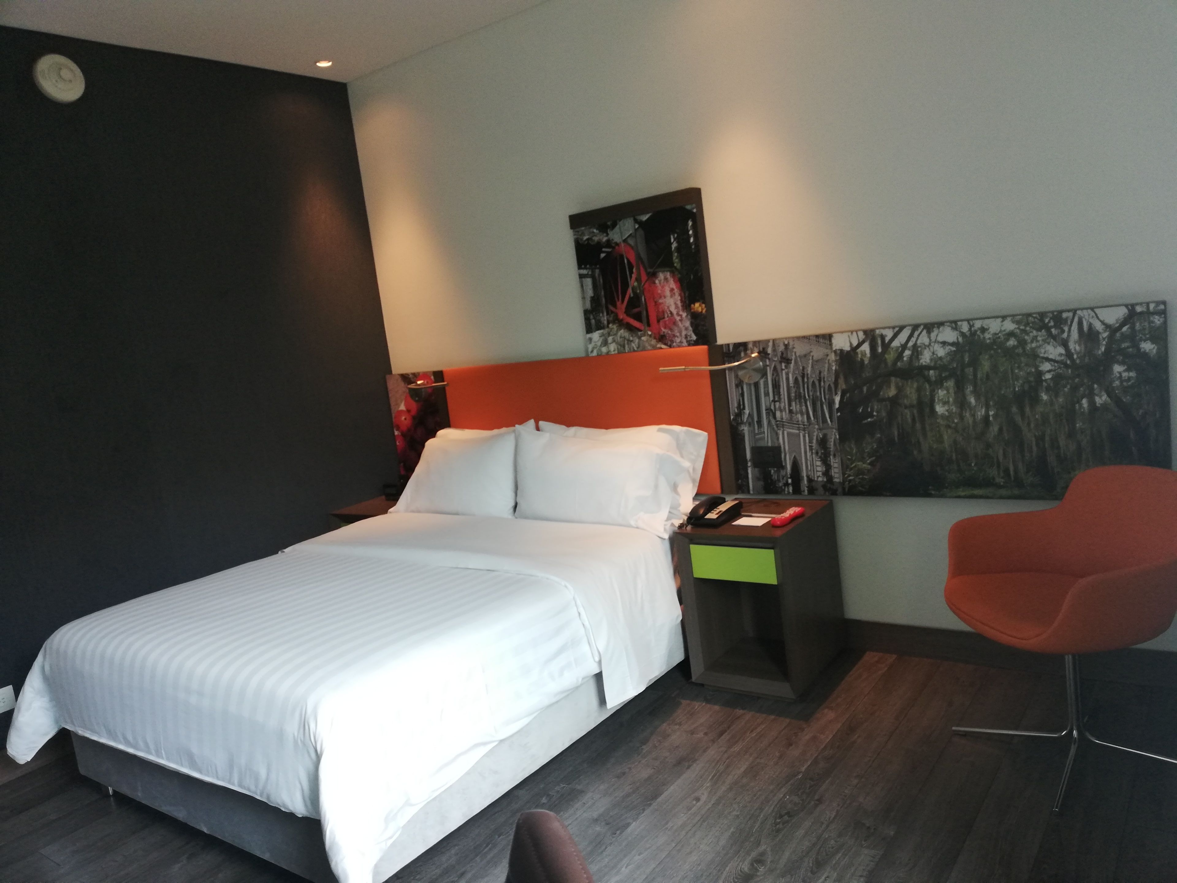 A modern hotel room with a large bed, a nightstand, a red chair, and a painting on the wall.