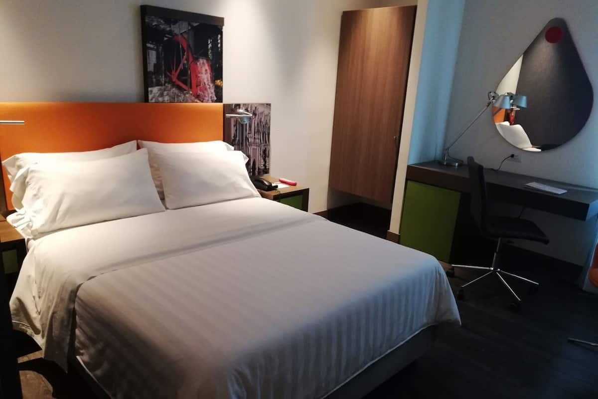 A hotel room with a bed, a desk, a chair, a nightstand, and a wall-mounted lamp.