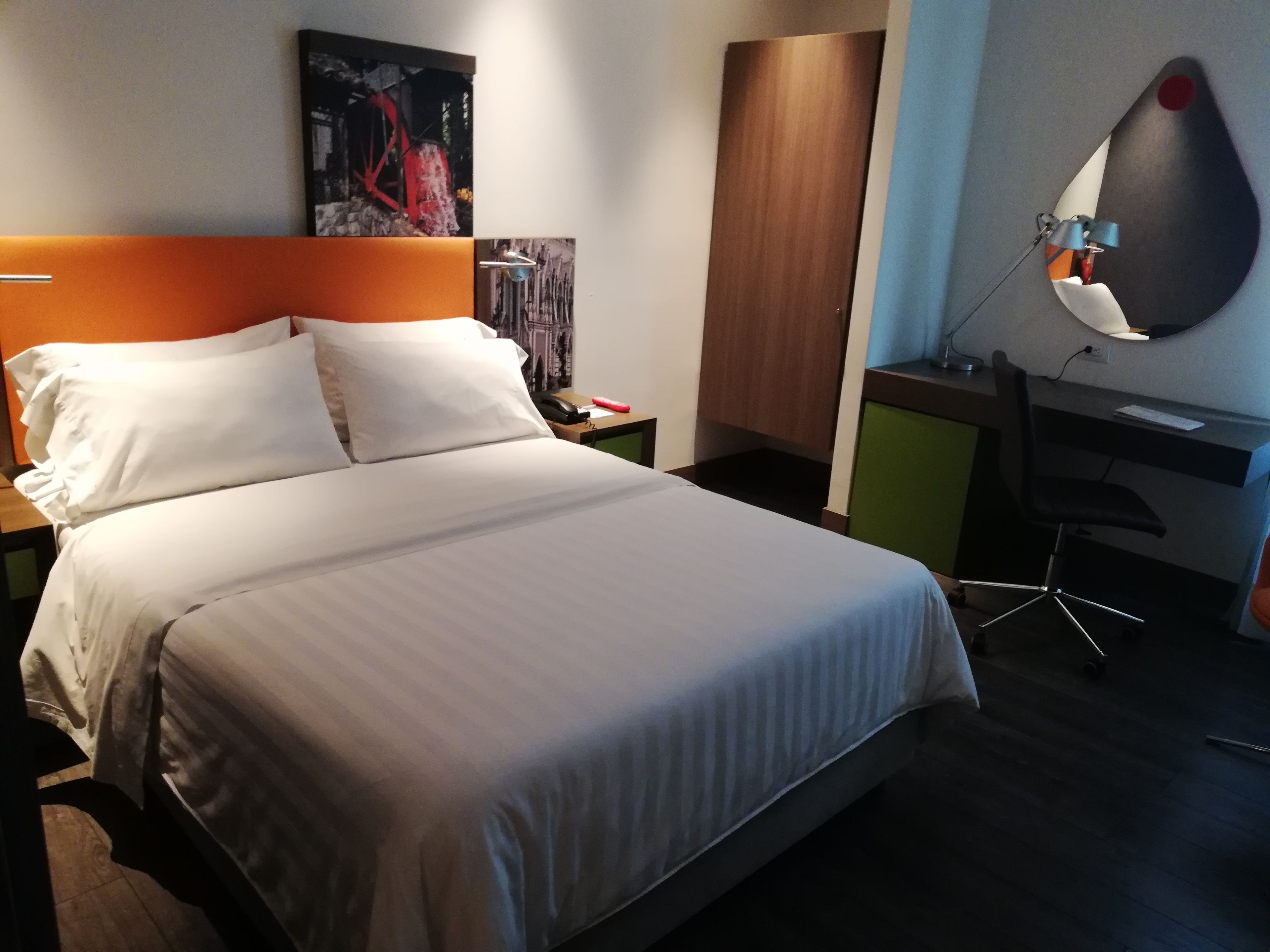 Ein ordentlich bezogenes Bett mit weißen Leinen, ein Holz-Kopfteil, ein Nachttisch mit Telefon und einem roten Gegenstand, ein Schreibtisch mit Stuhl und ein an der Wand befestigter Spiegel.