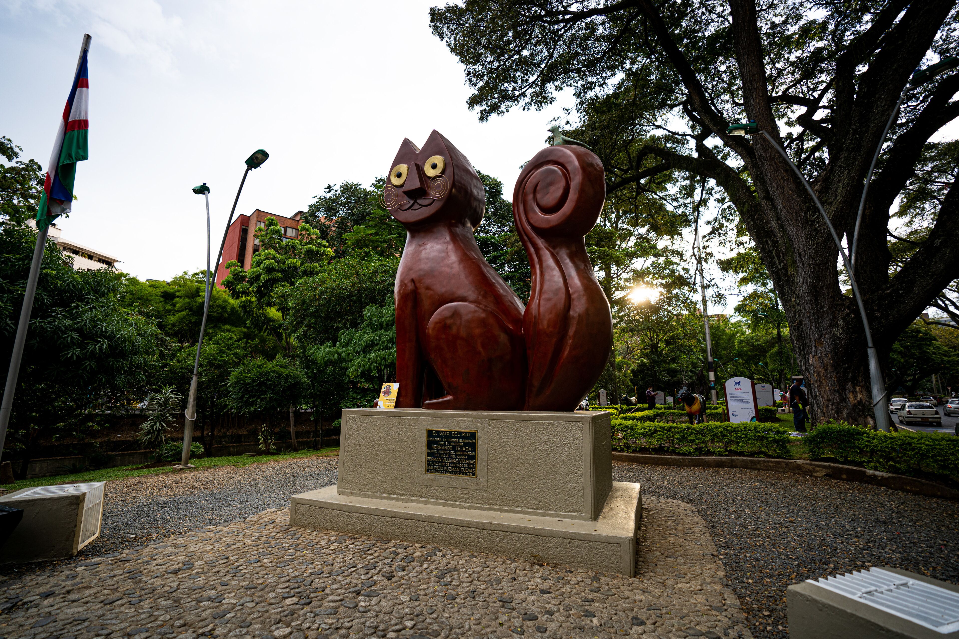 Una escultura de un gato en un parque público, con una placa, árboles y una bandera al fondo.