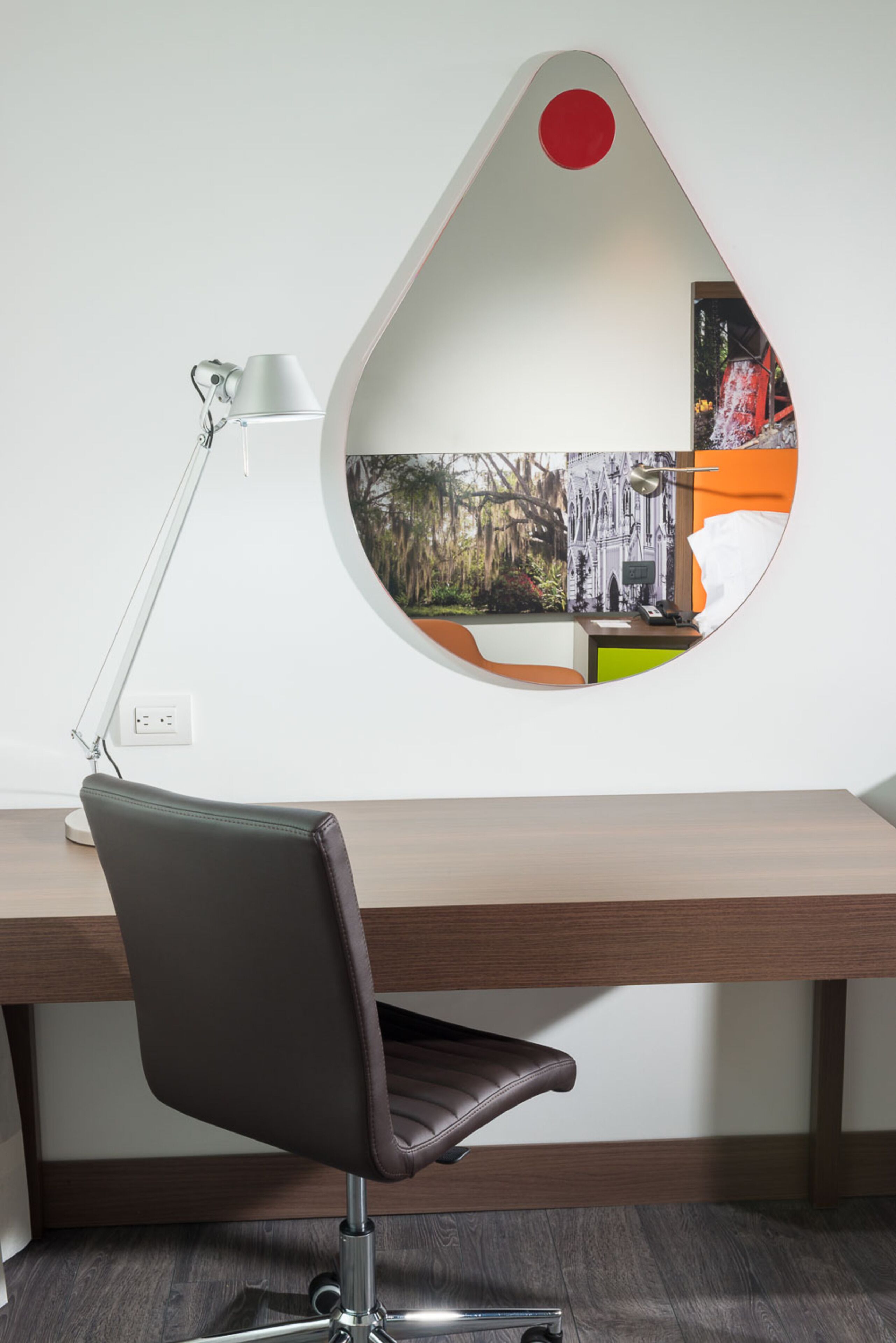 A modern office desk with a leather chair, a desk lamp, and a unique wall mirror reflecting a cityscape.