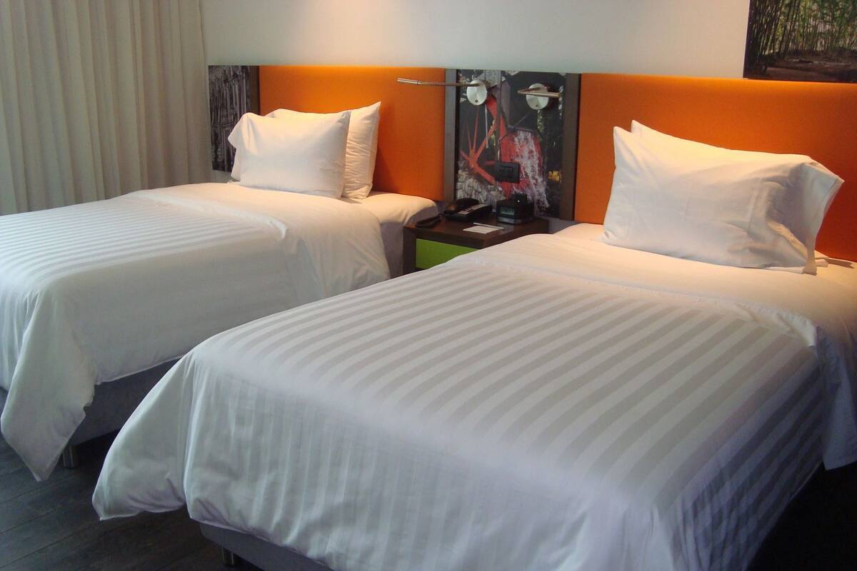 A hotel room with two beds, each with white bedding and pillows, a nightstand with a lamp, and a painting on the wall.