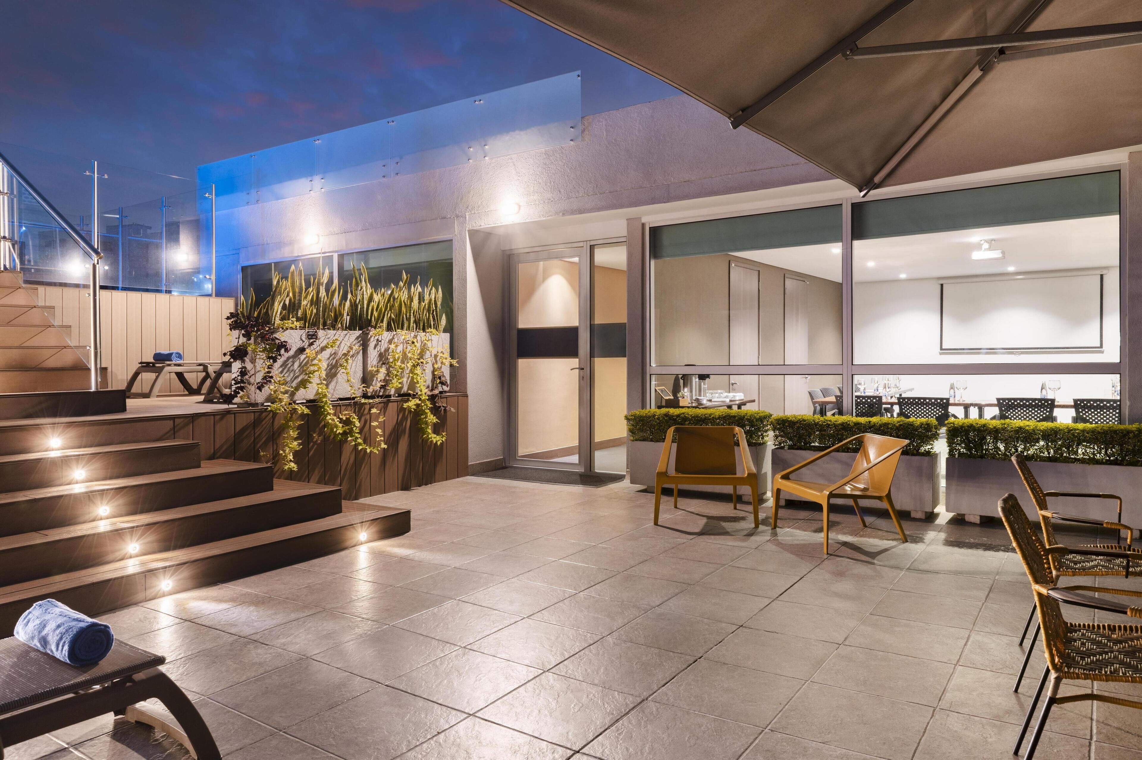 Un patio exterior moderno con una escalera que lleva a un nivel superior, una zona de estar con sillas y una barra.