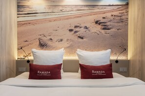 Un lit avec deux oreillers portant l’inscription « RAMADA » et, au mur derrière, une image représentant une plage.