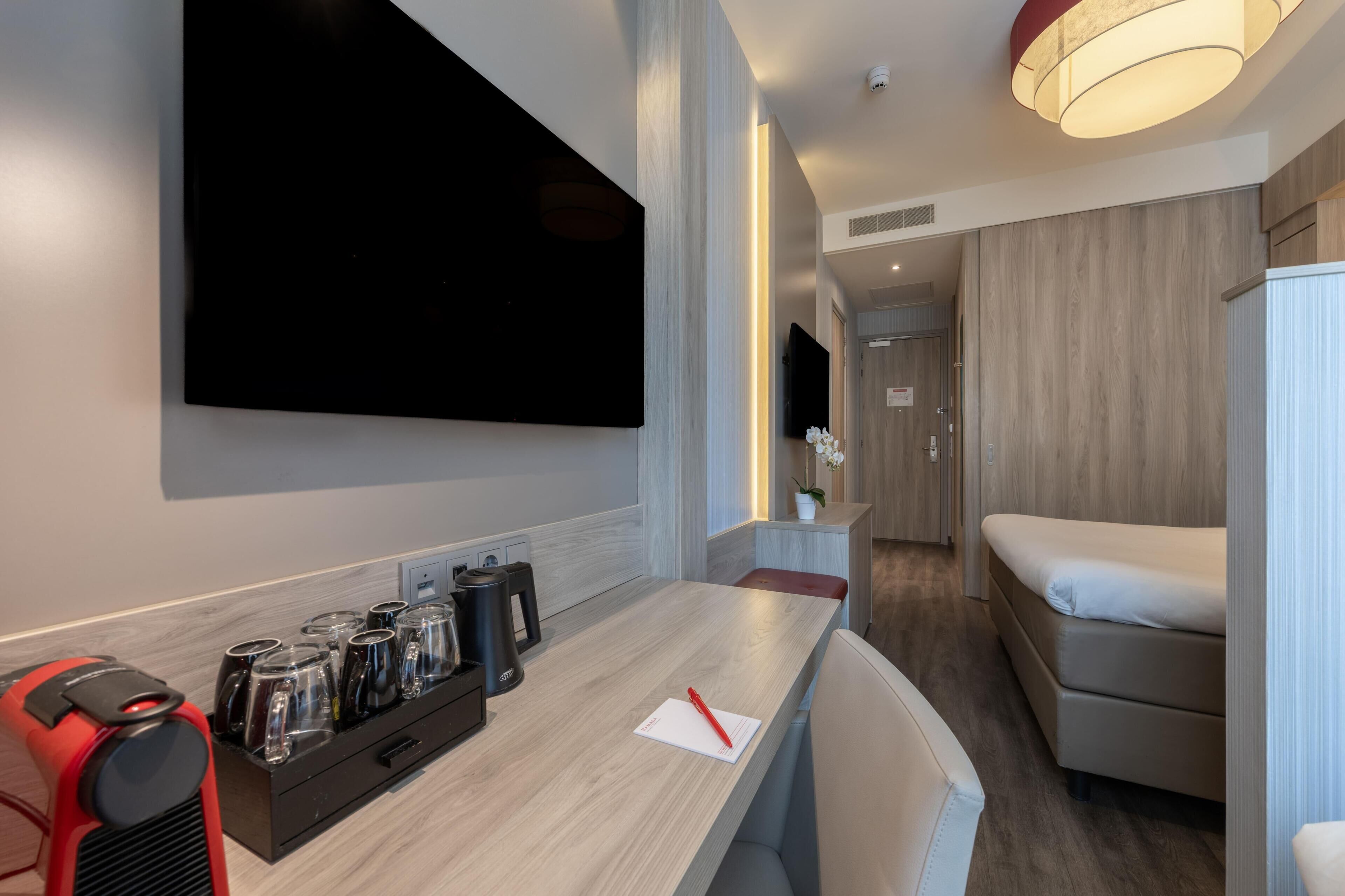 A modern hotel room with a flat-screen TV mounted on the wall, a wooden dining table with a coffee maker, a sofa, and a small seating area.