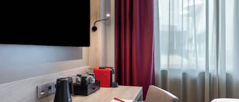 A hotel room with a wooden desk, a flat-screen TV mounted on the wall, a red suitcase, a black coffee maker, and a bed with white bedding.