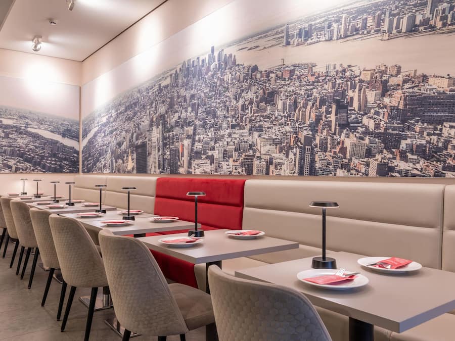 Un restaurante con un mural de la ciudad, mesas con cabinas y sillas, y platos con cubiertos.