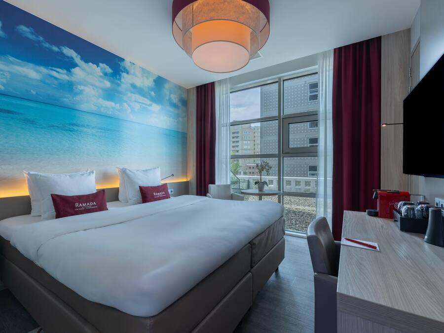 A hotel room with a bed, a desk, a TV, and a view of the city through large windows.