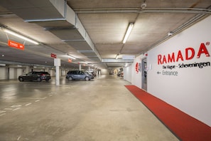 Eine Parkgarage mit mehreren parkenden Autos, Schildern mit der Aufschrift 'RAMADA' und 'Einfahrt' sowie einem roten Teppich, der zur Einfahrt führt.