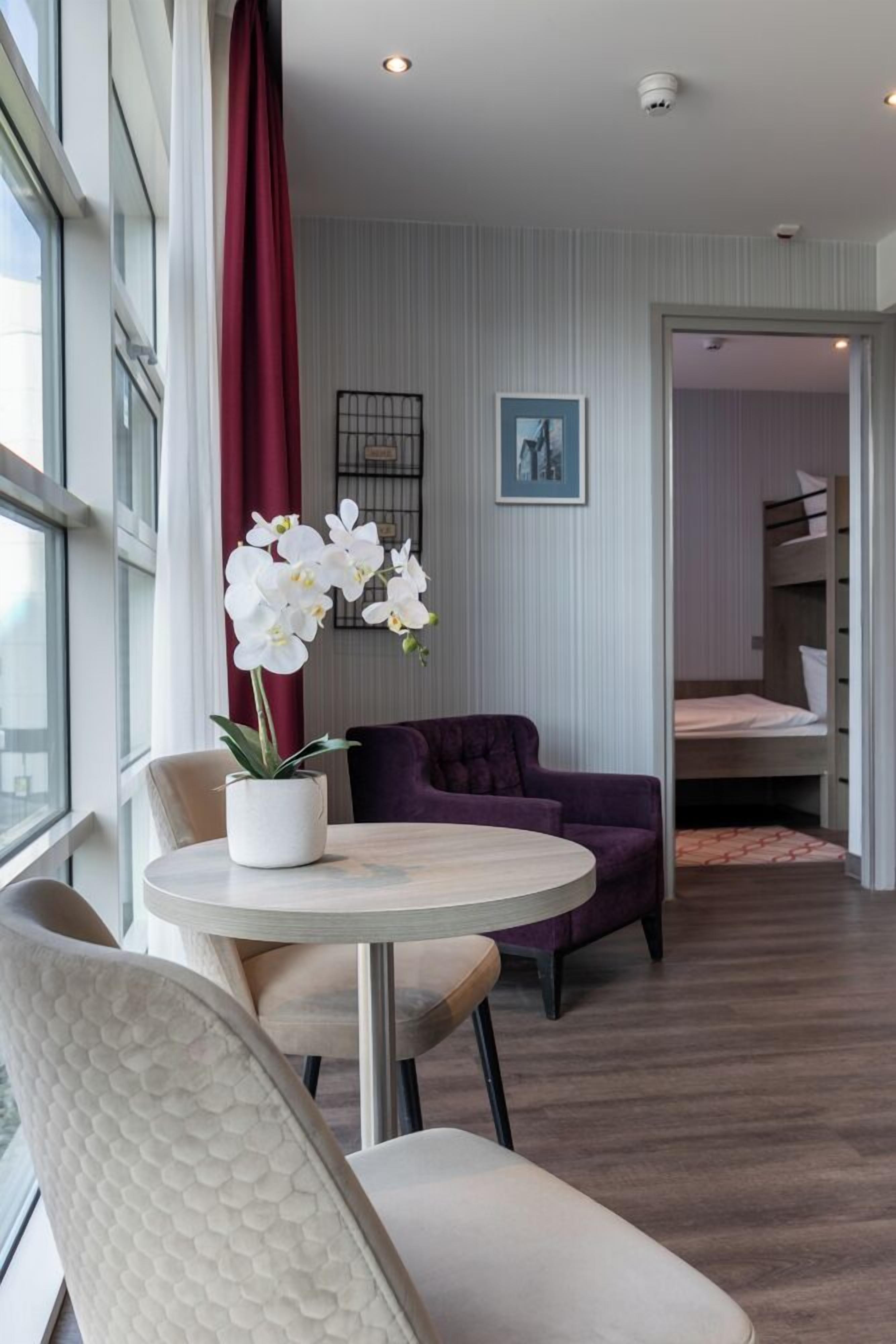 A hotel room with a round table, a purple sofa, a potted orchid, and a bunk bed in the background.