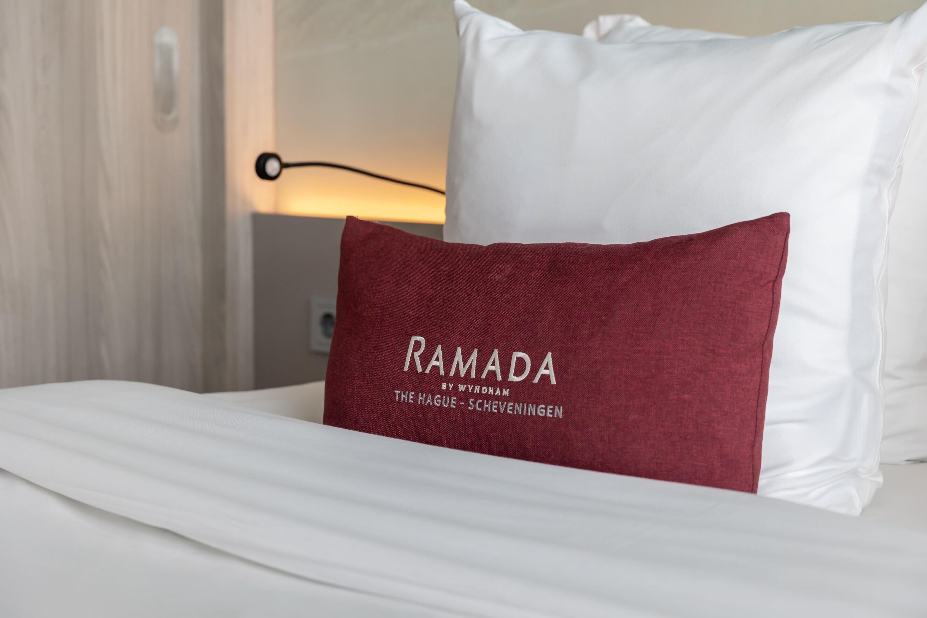 Ein Bett mit weißer Bettwäsche und einem roten Kopfkissen mit dem Aufdruck 'RAMADA BY WYNDHAM THE HAGUE - SCHEVENINGEN'.