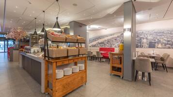 Een modern café met een buffet, een zithoek en een stadsgezicht muurschildering.