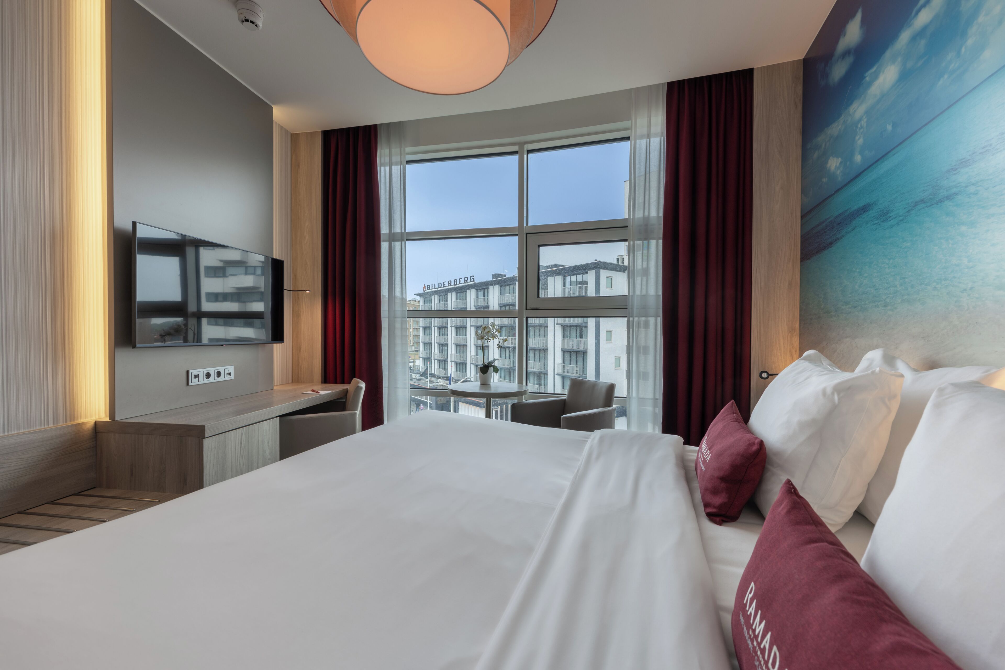 Ein Hotelzimmer mit Bett, Schreibtisch, Fernseher und einem großen Fenster mit Blick auf Gebäude.