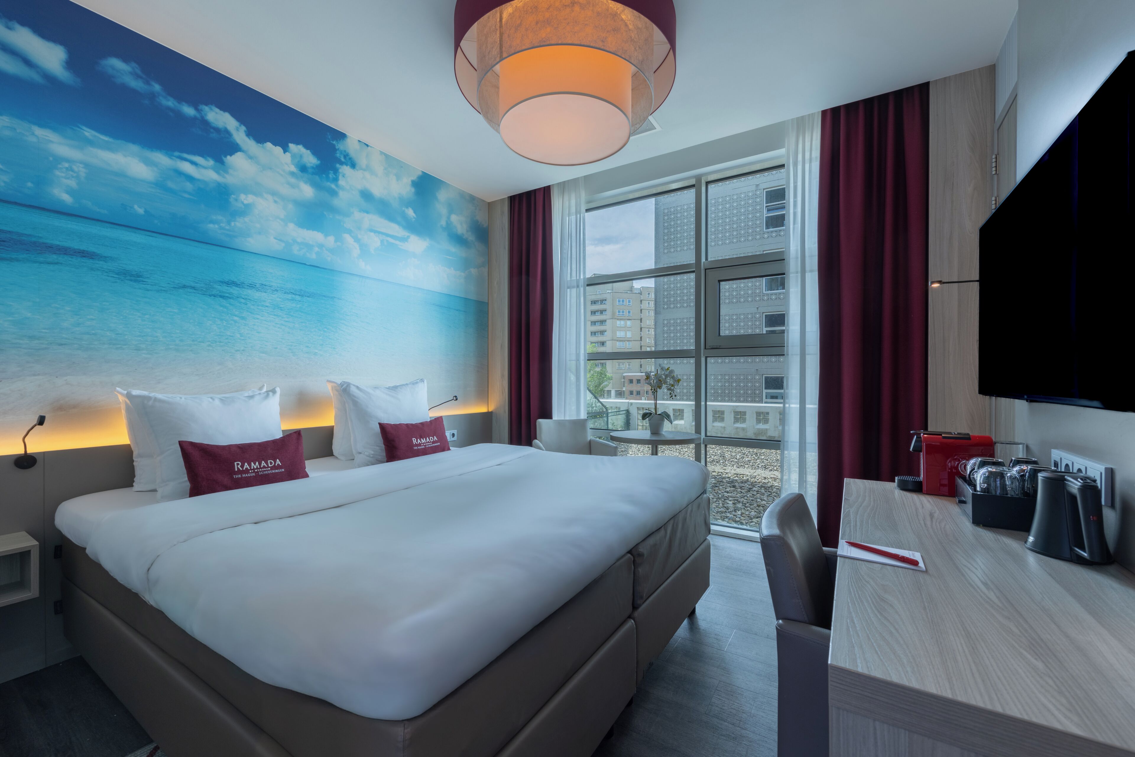 Habitació d'hotel amb llit, escriptori, televisió i vistes a la ciutat a través de grans finestres.