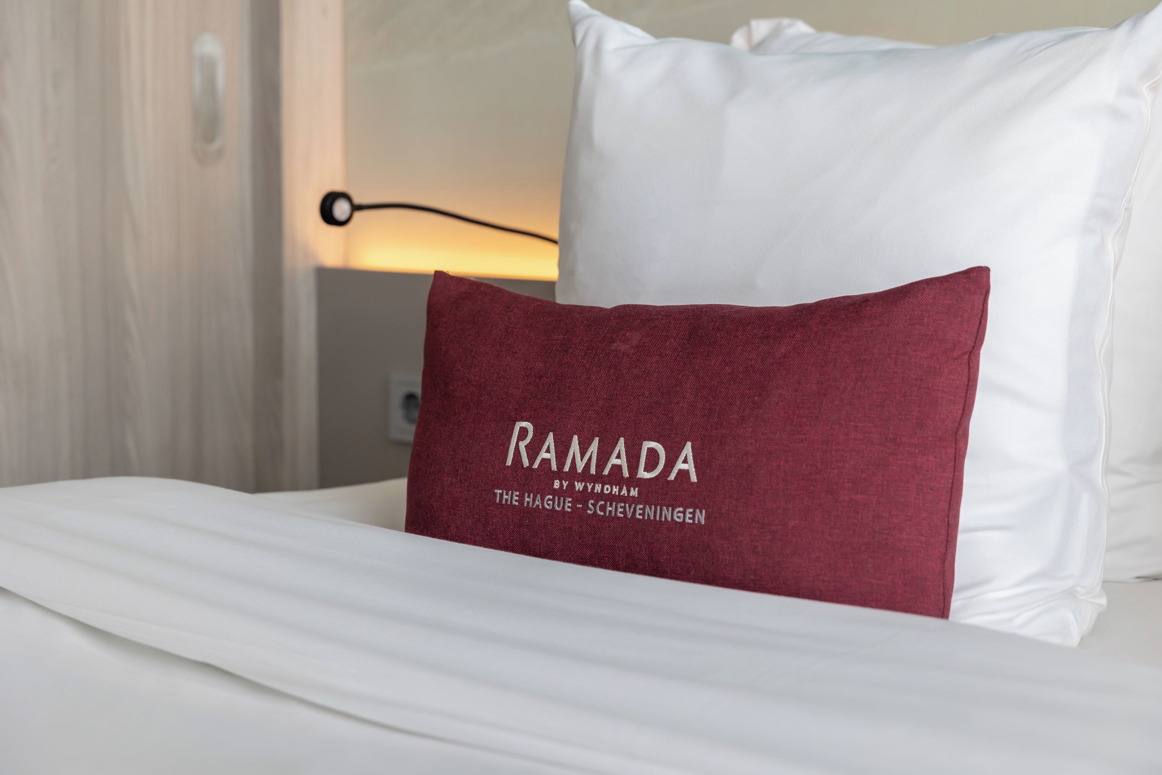 En säng med vita sängkläder och en röd kudde med texten ”RAMADA BY WYNDHAM THE HAGUE – SCHEVENINGEN”.