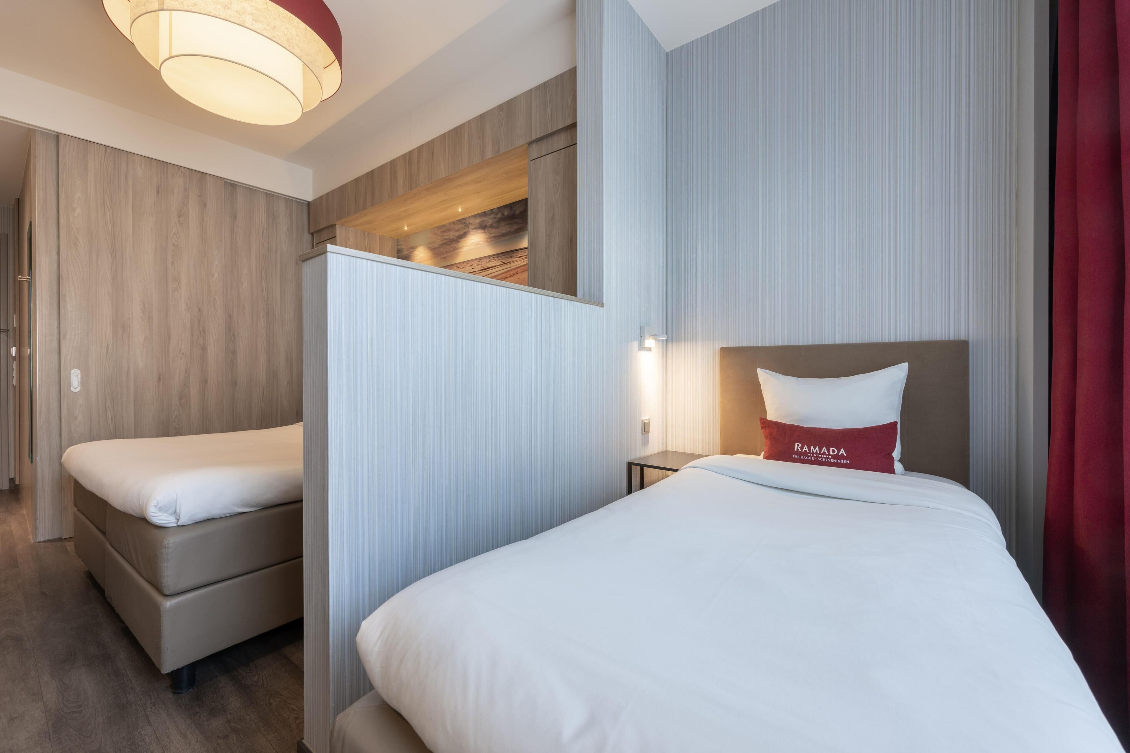 Habitación de hotel con dos camas, una mesita de noche, una lámpara de pared y vistas a la playa a través de una ventana.