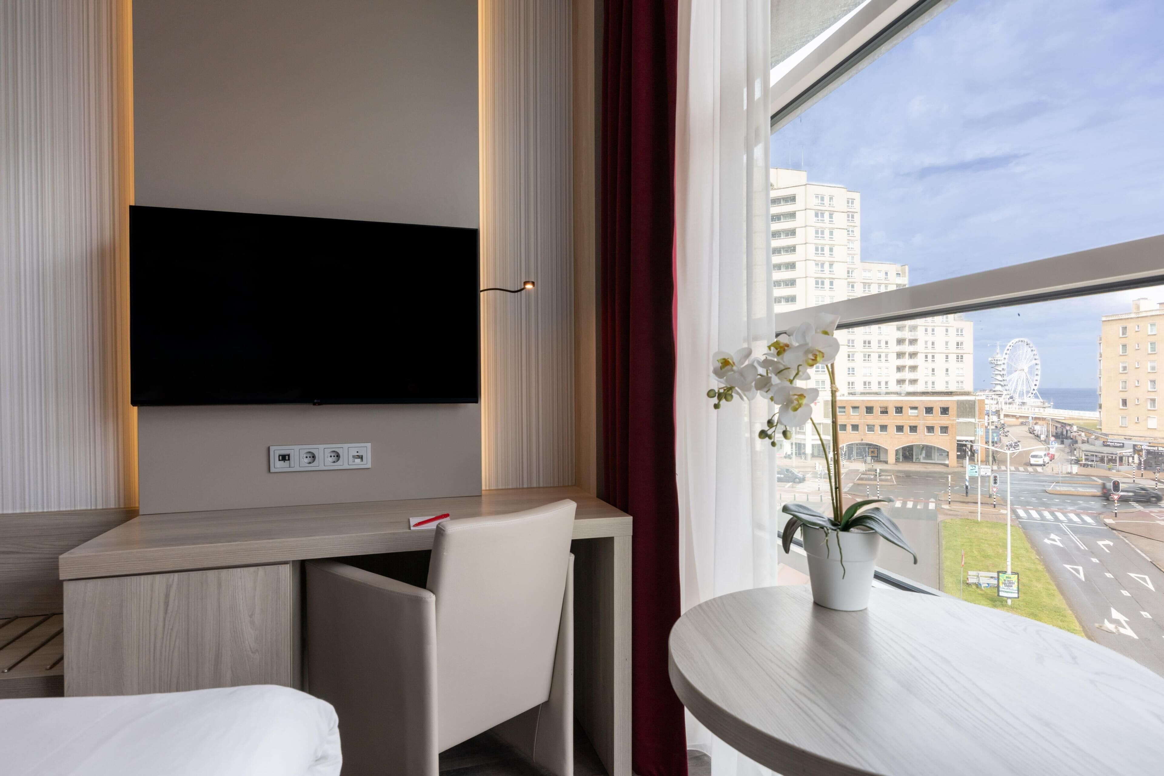 Une chambre d’hôtel équipée d’un téléviseur à écran plat fixé au mur, d’un bureau avec une chaise, d’une petite table avec un pot de plante et d’une fenêtre donnant sur la ville.