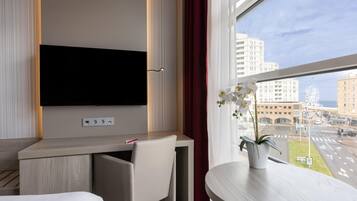 Une chambre d’hôtel équipée d’un téléviseur à écran plat fixé au mur, d’un bureau avec une chaise, d’une petite table avec un pot de plante et d’une fenêtre donnant sur la ville.