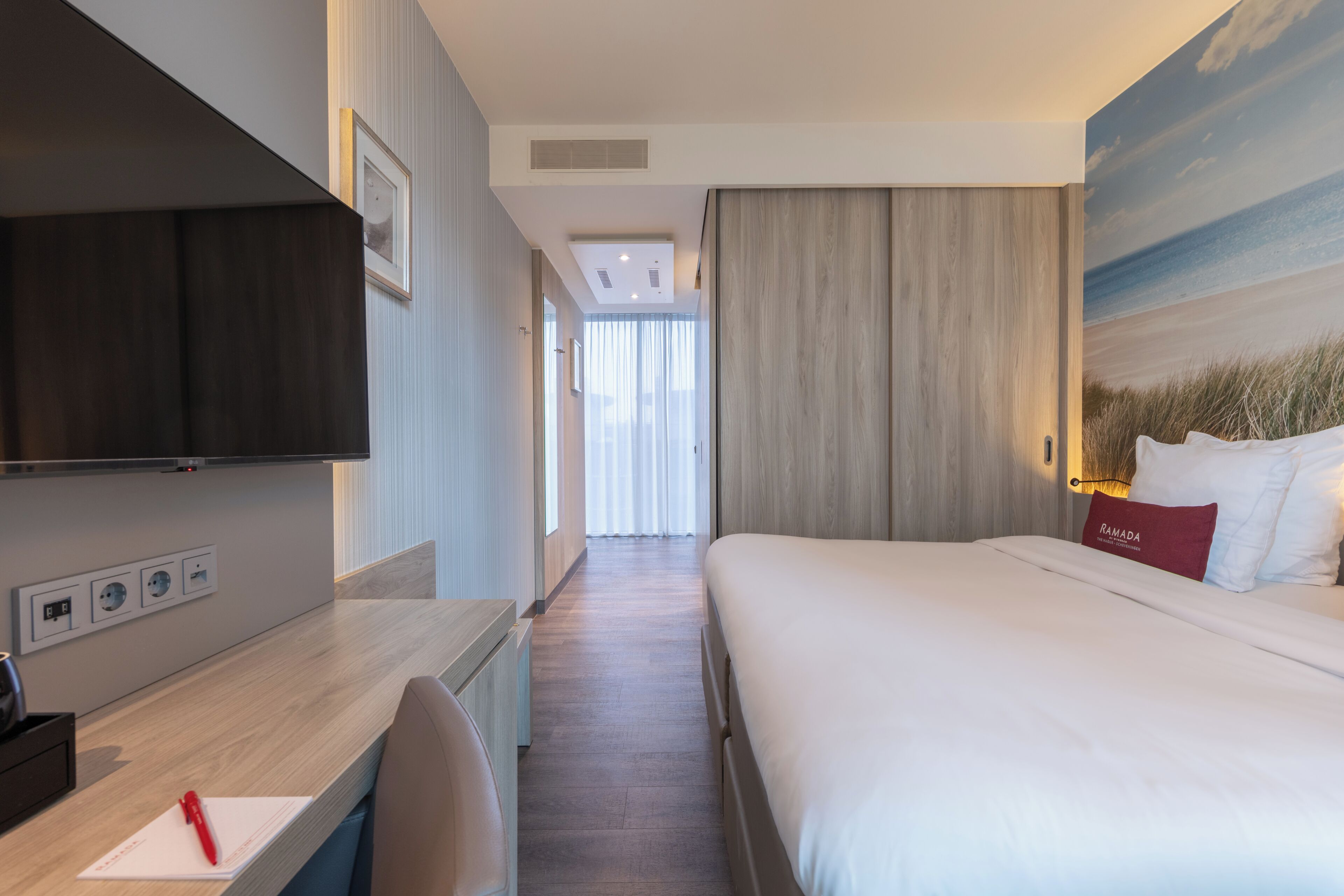 Une chambre d’hôtel avec un lit, un bureau, une chaise, une télévision et un tableau représentant une plage.