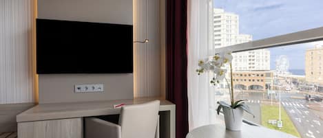Habitación de hotel con televisor de pantalla plana montado en la pared, un escritorio con una silla, una orquídea en maceta en el alféizar de la ventana y vistas a la ciudad con una noria.