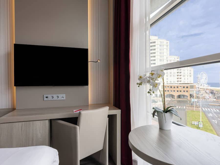 A hotel room with a flat-screen TV mounted on the wall, a desk with a chair, a potted orchid on the windowsill, and a view of the cityscape and a Ferris wheel.