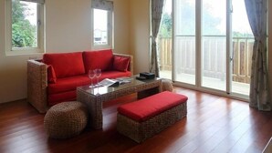 A spacious living room with a red sofa, a glass coffee table, and wicker ottomans. There are two windows with curtains, and a sliding glass door leading to an outdoor area. - Jimei Homestay (Ji'an)