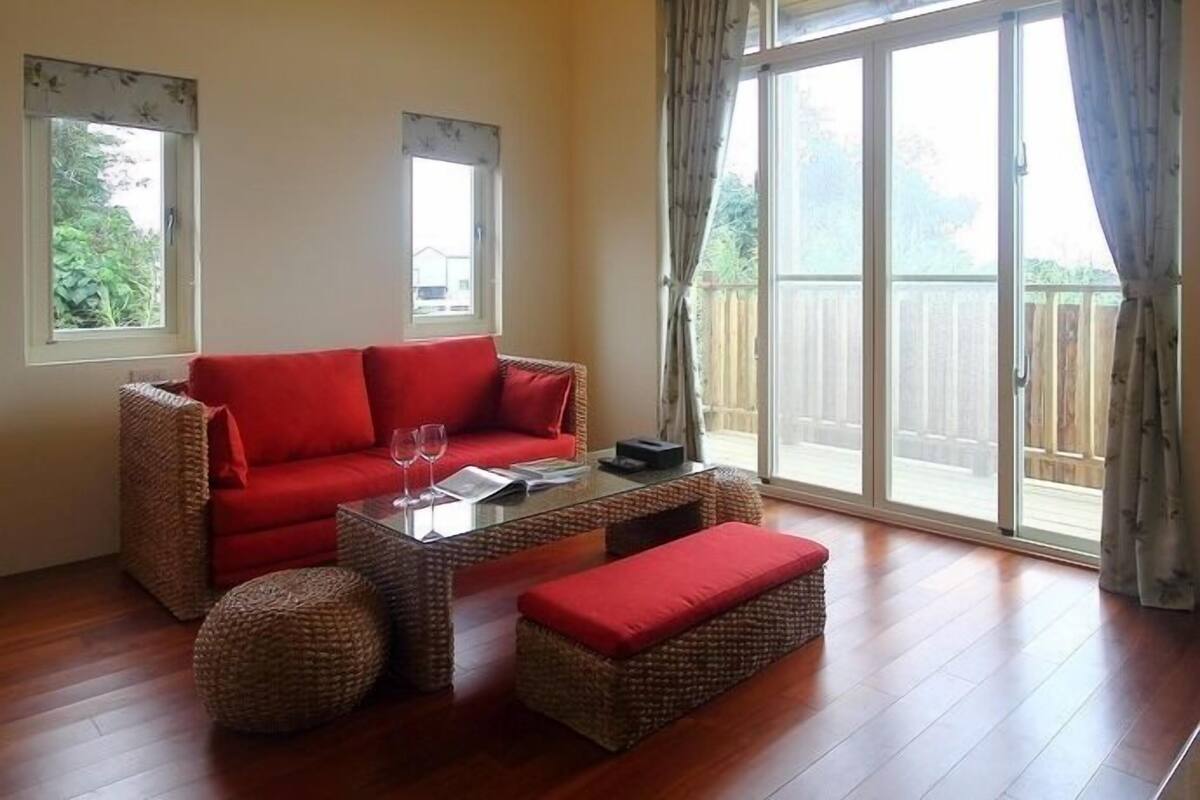 A spacious living room with a red sofa, a glass coffee table, and wicker ottomans. There are two windows with curtains, and a sliding glass door leading to an outdoor area.