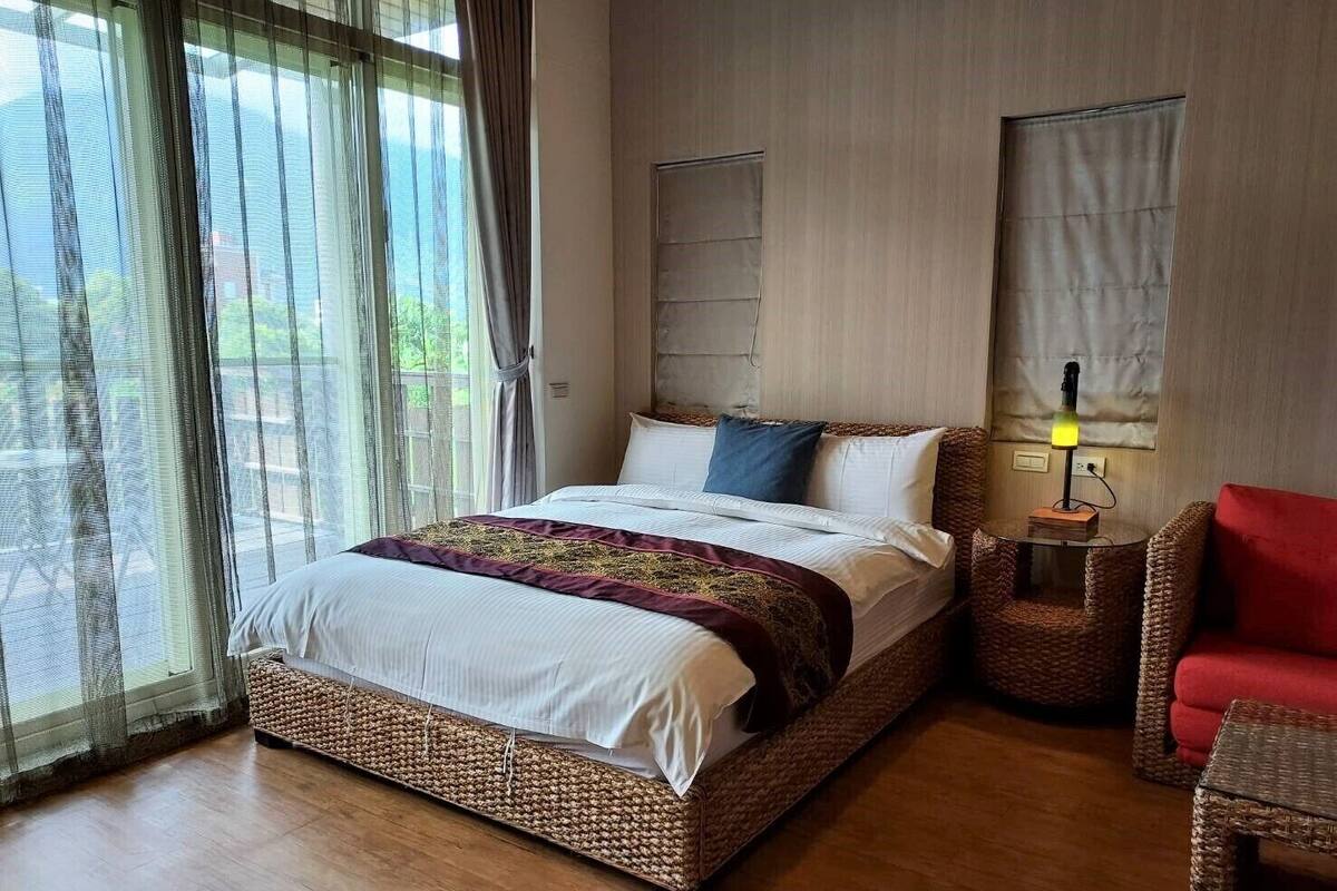 A neatly arranged bedroom with a large bed, a bedside table with a lamp, and a wicker chair.
