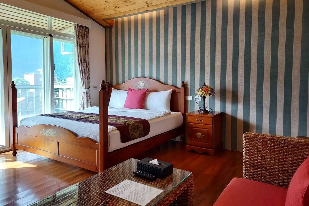 A bedroom with a four-poster bed, a nightstand with a lamp, a wicker chair, and a view of the sea through large windows.