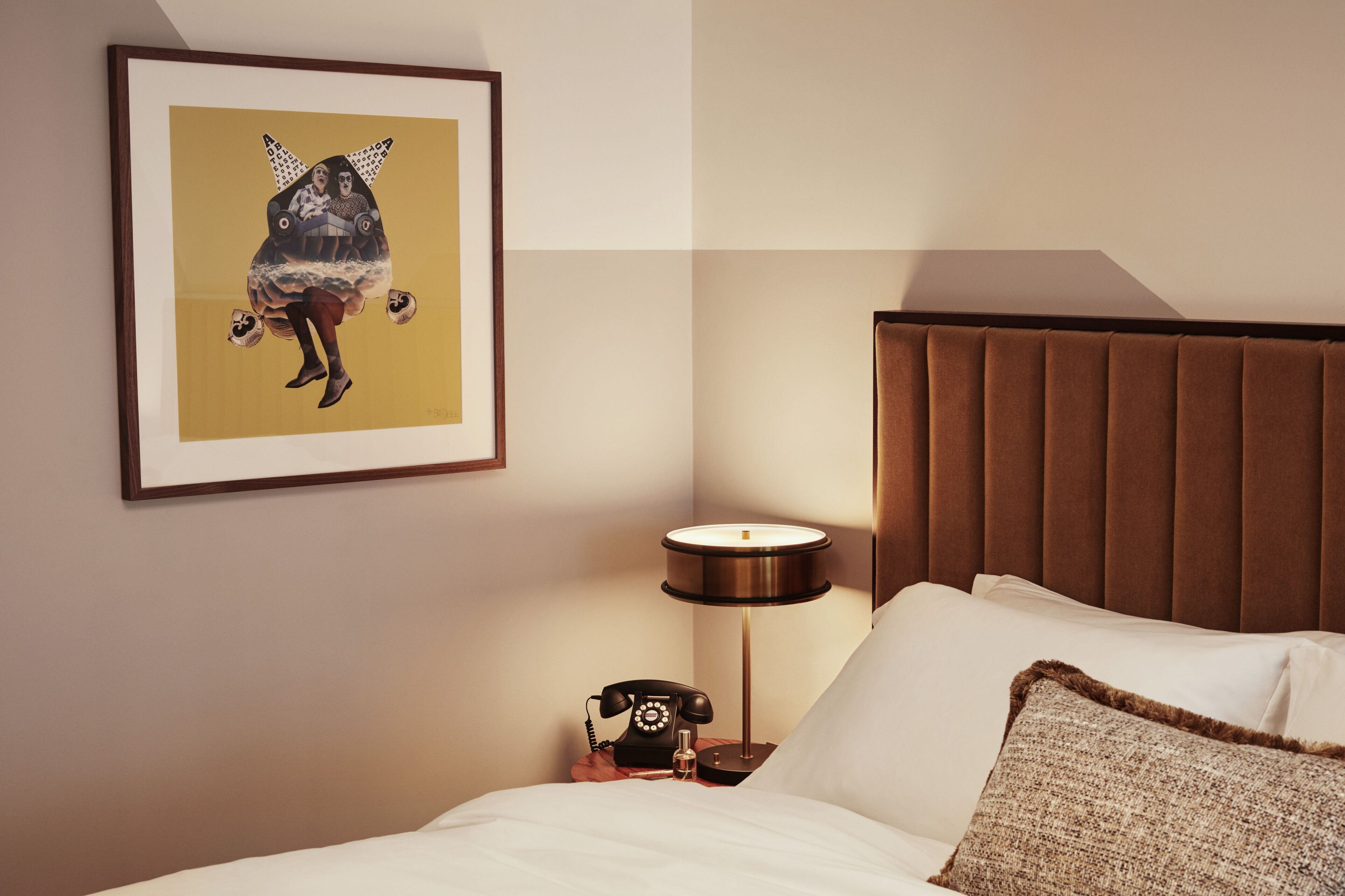 Una camera d'albergo con un letto, un comodino con una lampada, un quadro incorniciato e un poggiatesta marrone.