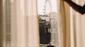 A table with a pair of glasses and a smartphone, curtains partially drawn, and a view of a Ferris wheel in the background. - The Hoxton, Holborn (London)