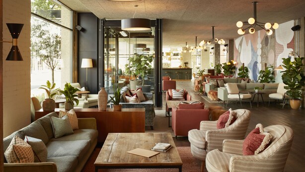 A spacious lobby with a wooden coffee table, sofas, and armchairs. There are potted plants and a large window providing natural light. - The Hoxton, Holborn (London)