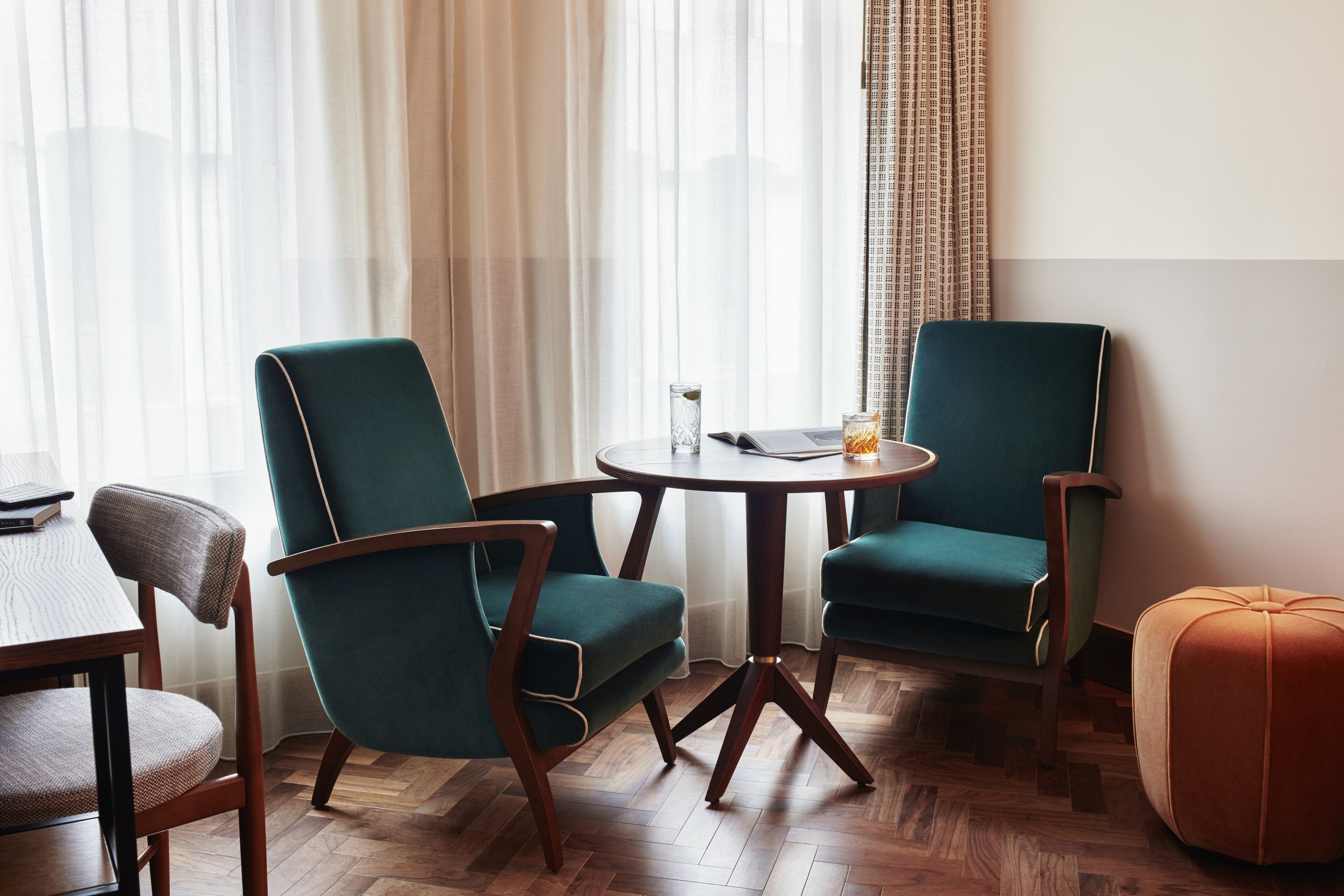 A room with two teal armchairs, a wooden dining table, and a small round table with a glass and a book.