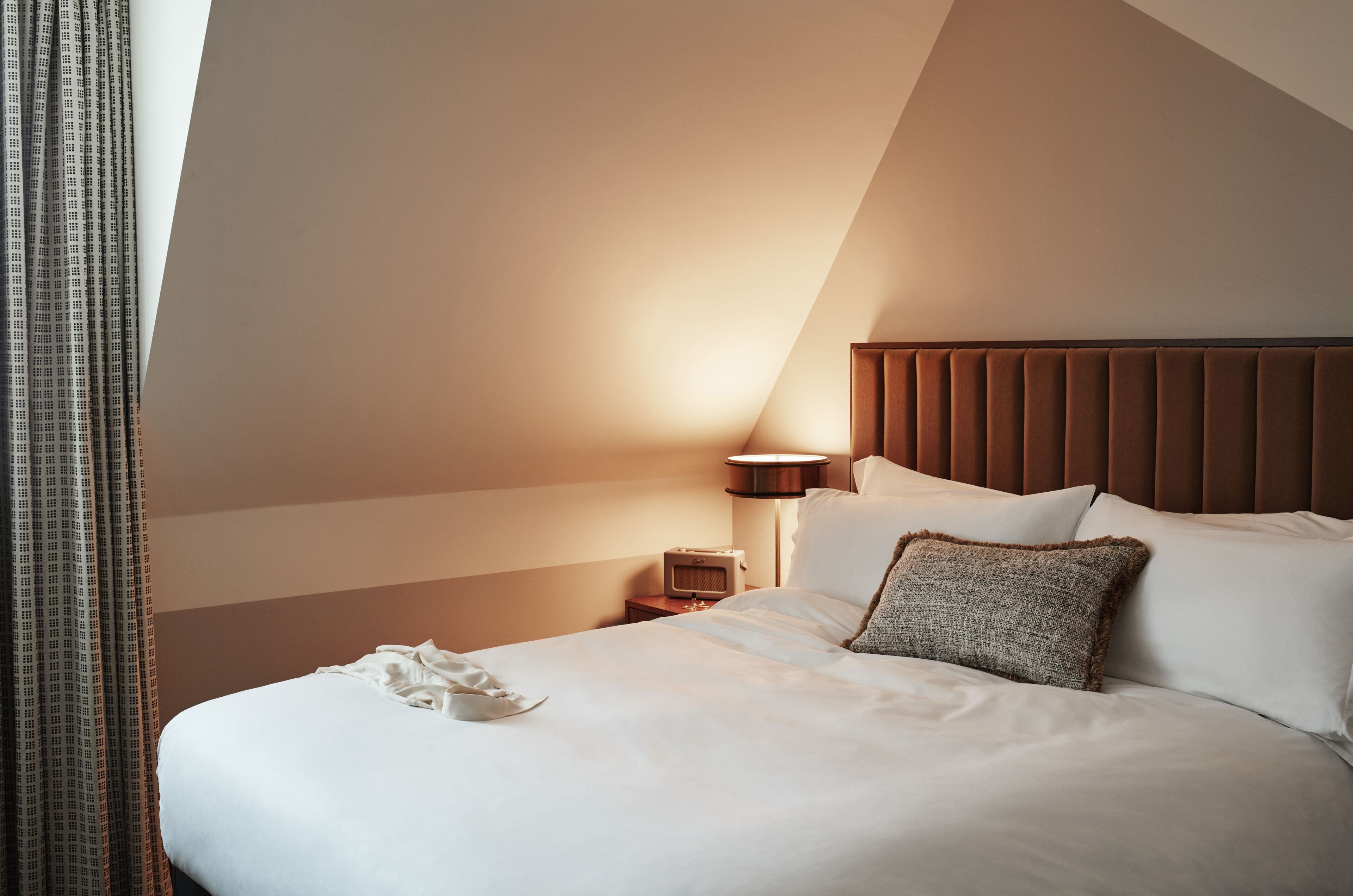 Ein Hotelzimmer mit einem großen Bett, einem Nachttisch mit Lampe und einem Fenster mit Vorhängen.