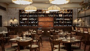 A restaurant with wooden tables and chairs, a bar area with shelves of bottles, and pendant lighting. - The Hoxton, Holborn (London)