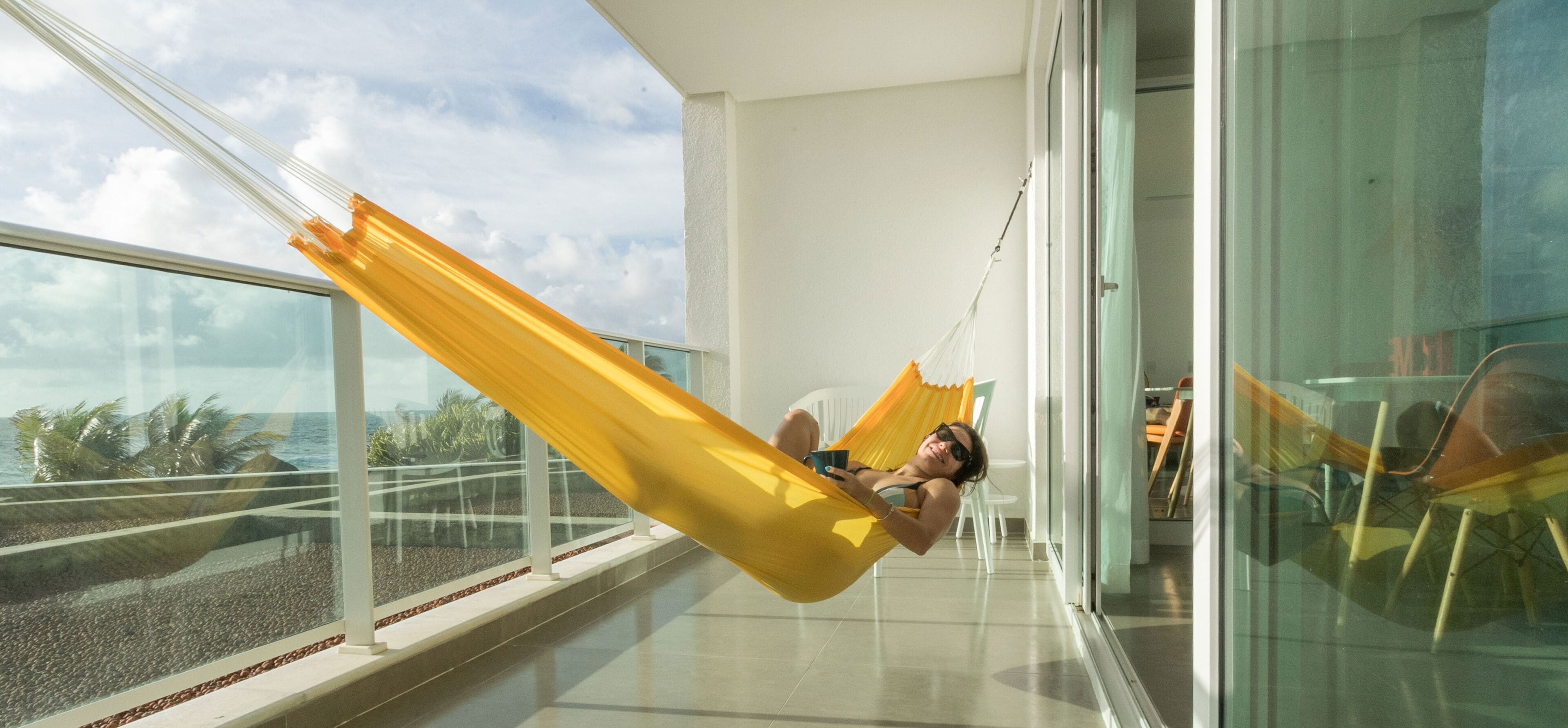 Una persona descansando en una hamaca amarilla en un balcón con vistas a la playa y palmeras.