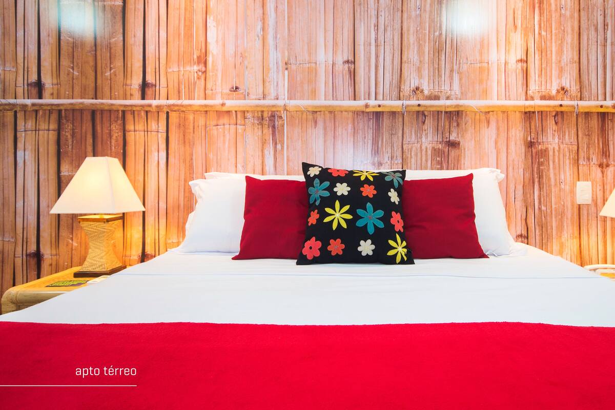 Ein Bett mit weißer Bettwäsche, einer roten Decke und einem schwarzen Kopfkissen mit bunten Blumen. Auf beiden Seiten des Bettes stehen jeweils Nachttischlampen.