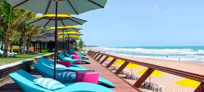 Un espace de détente aménagé au bord de la plage, avec des chaises longues et des parasols, une terrasse en bois et une vue sur l’océan.