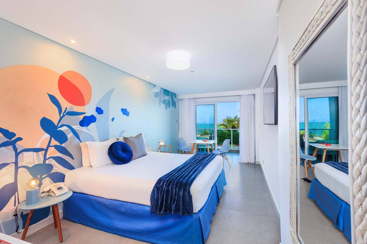 Ein modernes Hotelzimmer mit einem großen Bett, einem kleinen Tischchen mit Telefon und einem Balkon mit Blick auf den Ozean.