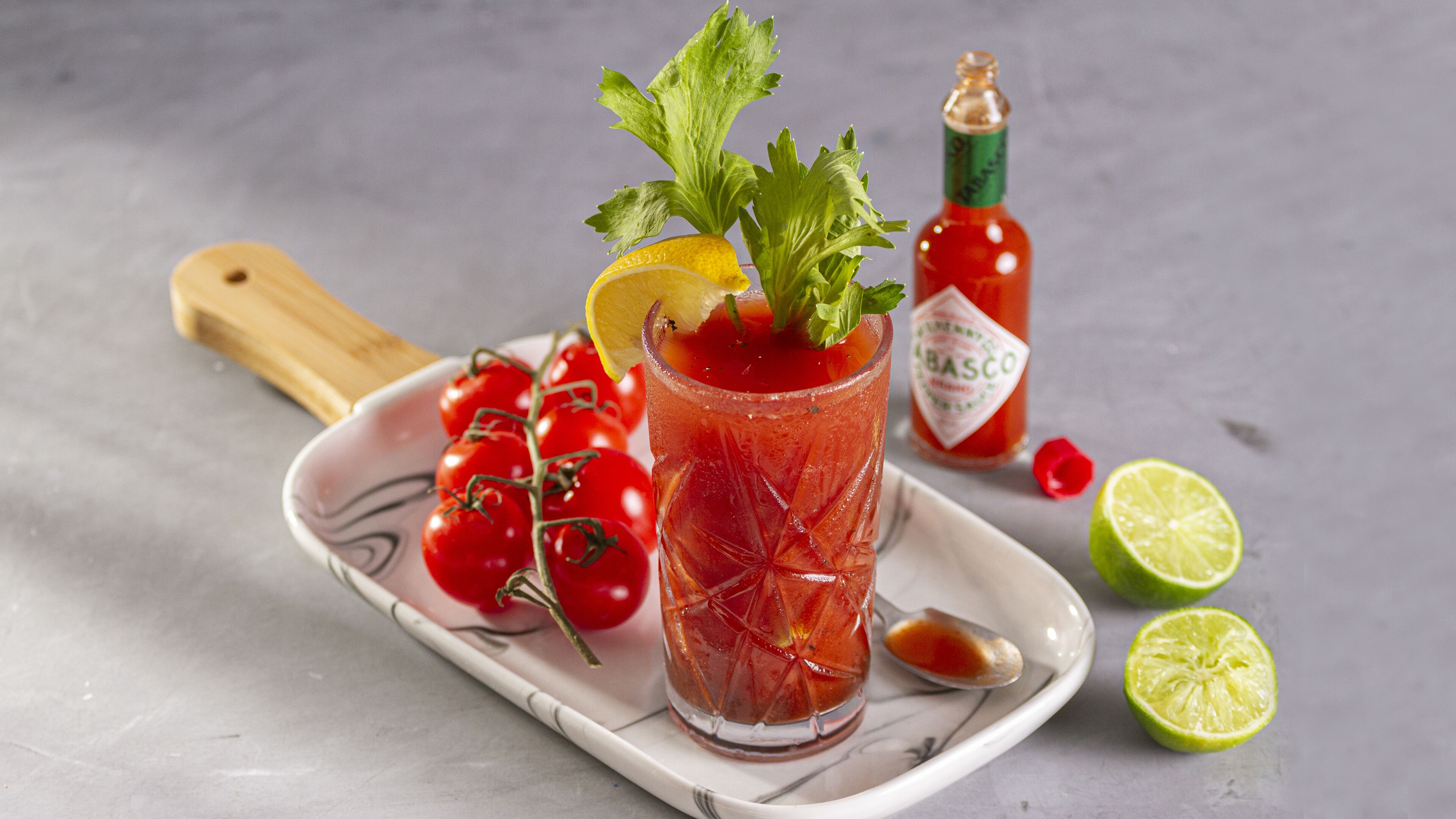 A glass of Bloody Mary with a celery stalk, lemon slice, and tomato garnish on a white plate. A bottle of Tabasco sauce and a wooden cutting board with tomatoes are also visible.