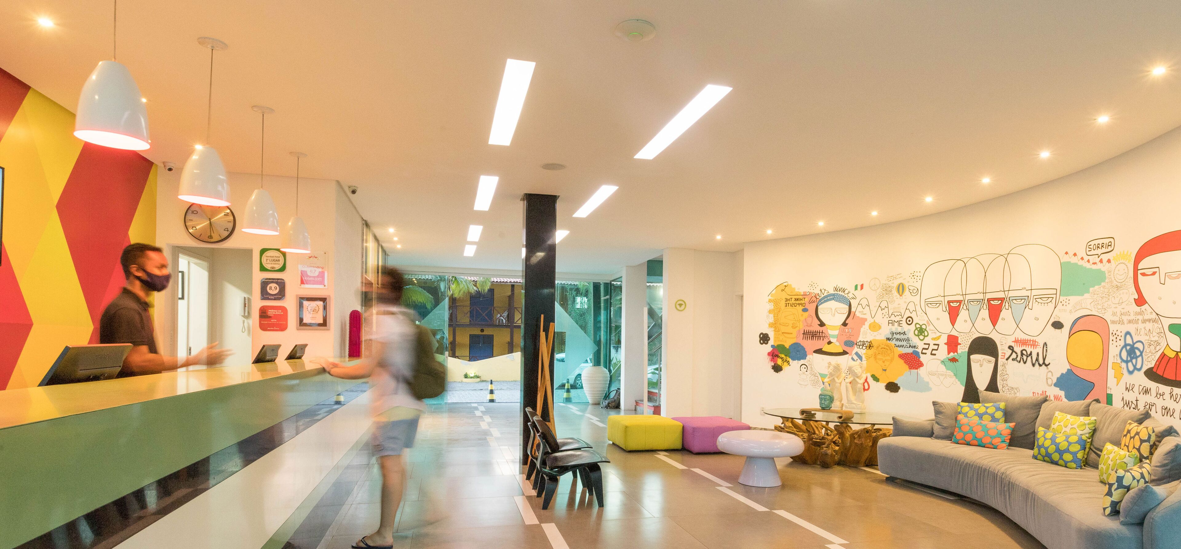 Een moderne hotellobby met een receptiebalie, kleurrijke muurkunst en een zithoek met een bank en voetenbankje.