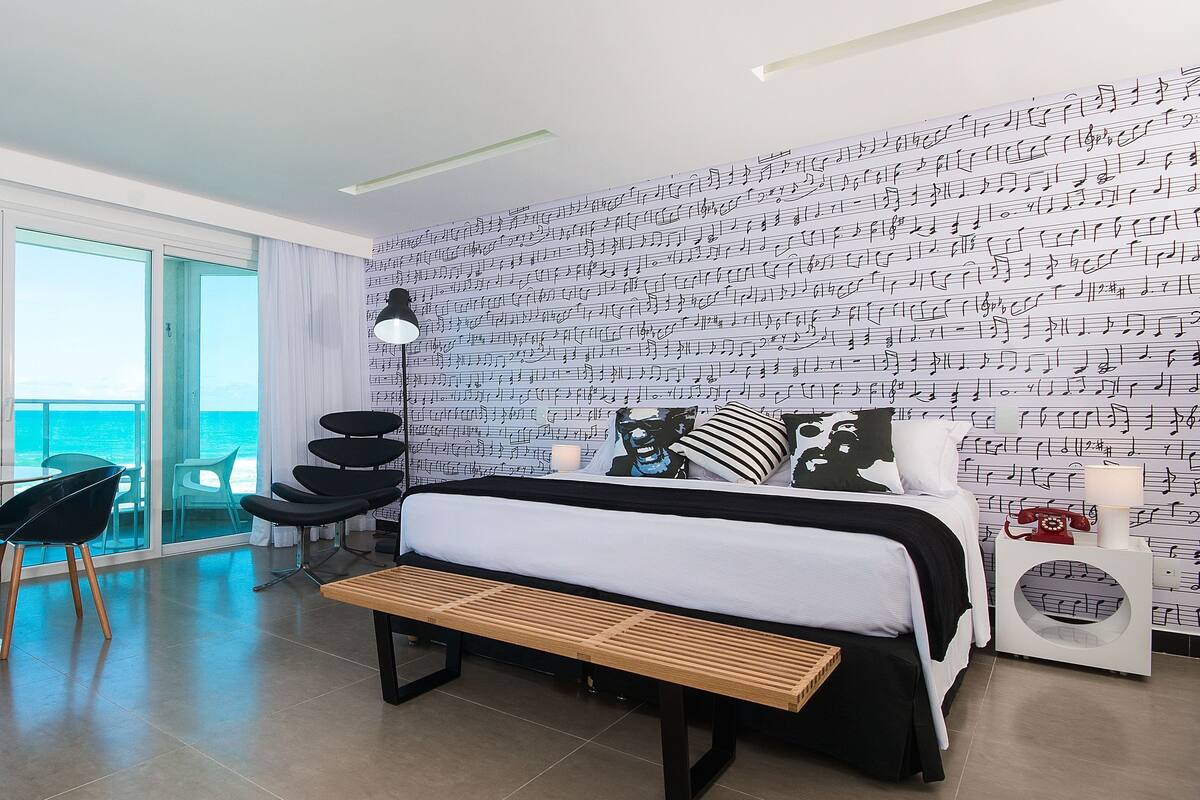 Ein modernes Hotelzimmer mit großem Balkon, einem Bett mit Kopfschild in Schwarz-Weiß-Muster und Blick auf den Ozean.
