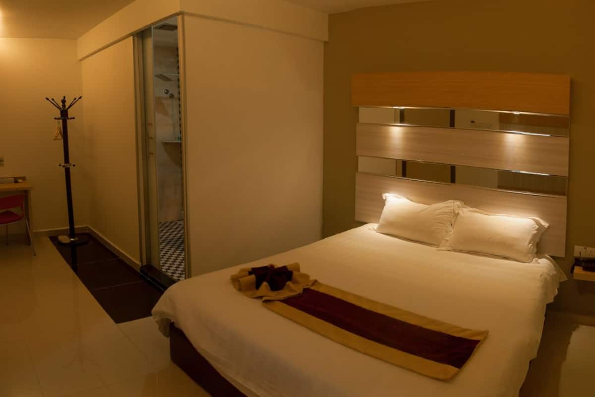 A hotel room with a bed, a coat rack, a shower, and a small table with a chair.