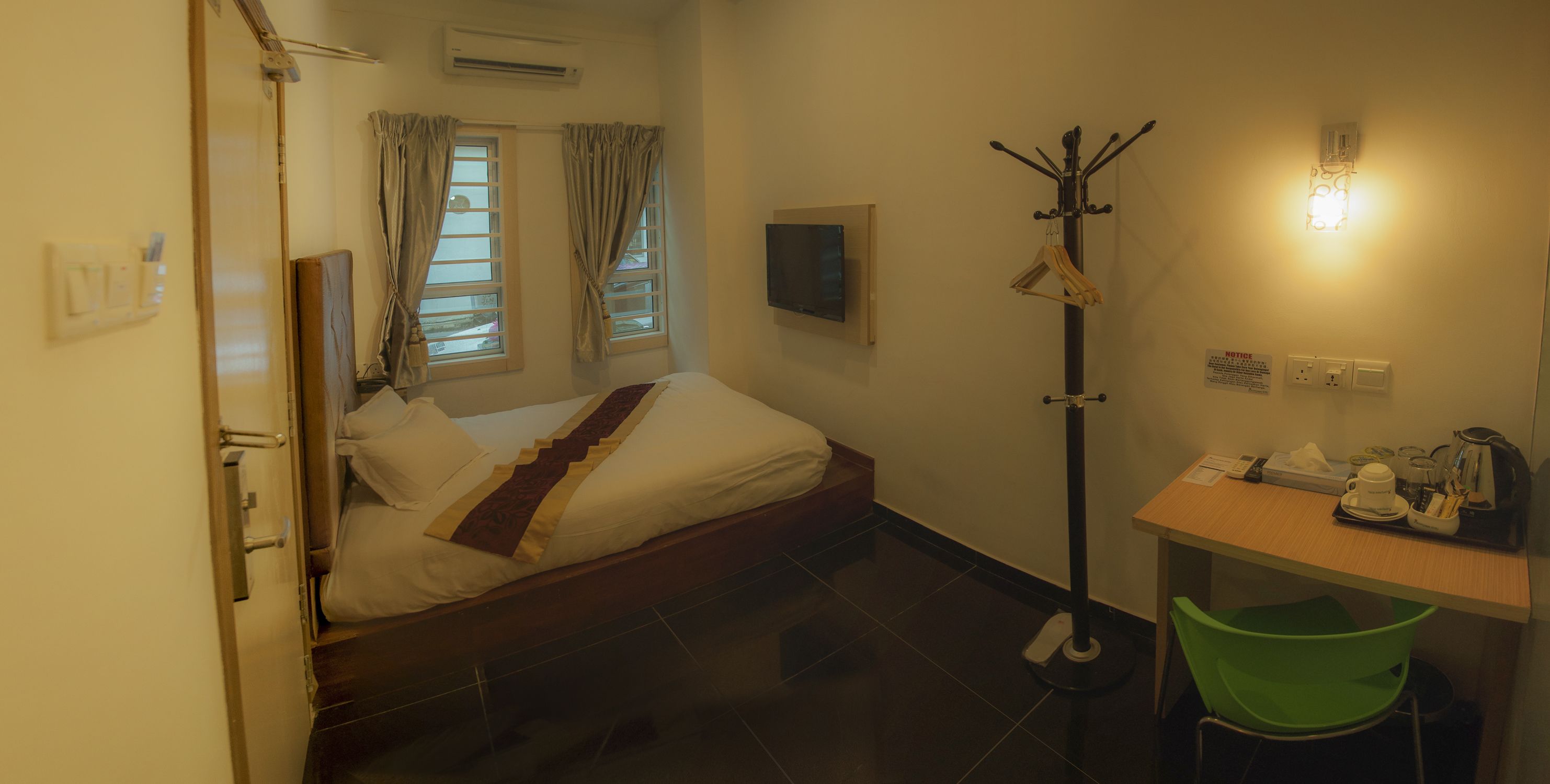 A hotel room with a bed, a desk with a chair, a coat rack, and a wall-mounted light.