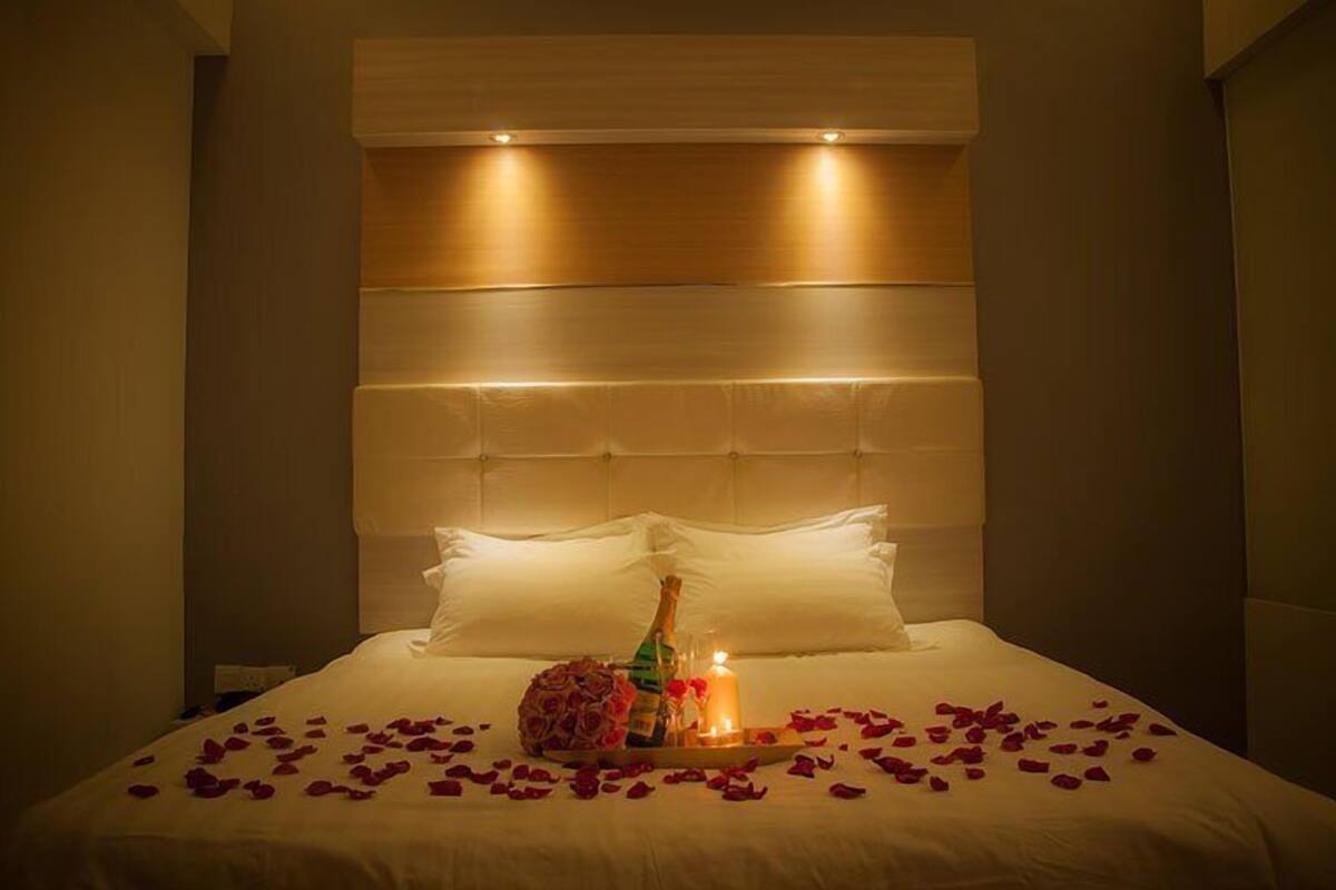 A hotel room with a large bed, a headboard, and a bedside table with a bottle, a candle, and rose petals.
