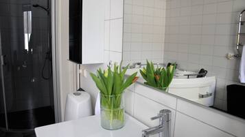A modern bathroom with a glass vase of green plants on the sink, a large mirror, and a shower area.