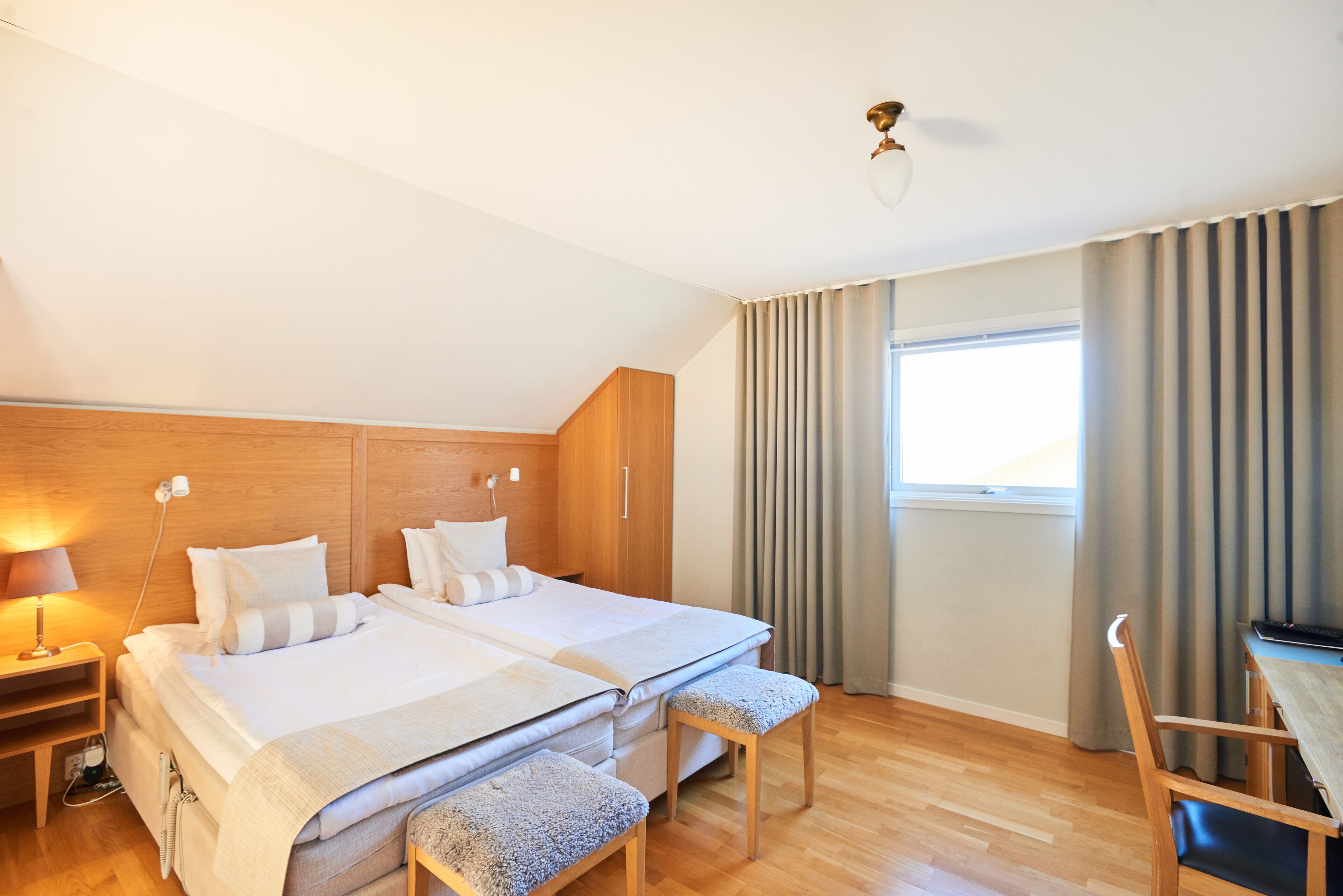 A hotel room with two beds, a desk, a chair, a lamp, and a window with curtains.