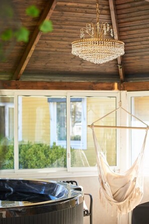 A wooden ceiling with a hanging chandelier, a hammock, and a hot tub in a room with large windows.